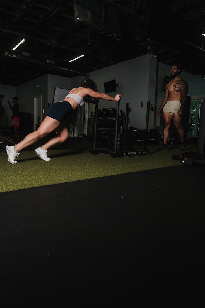 A muscular woman pushes workout equipment while a man observes in a gym setting.